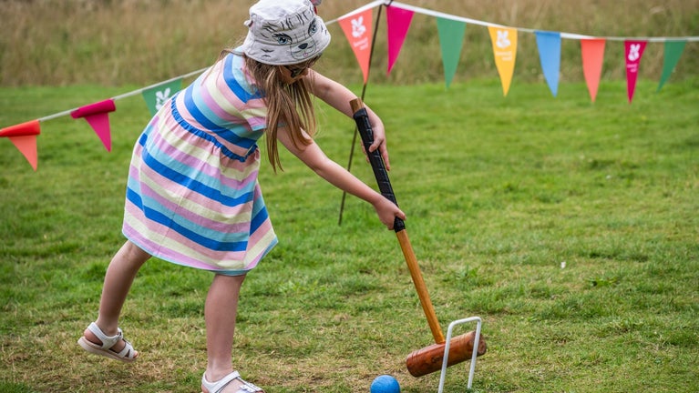 A young girl wearing a summer dress and hat is playing croquet in the lawns at Oxburgh Estate. Behind her you can see colourful bunting and Oxburgh Hall.